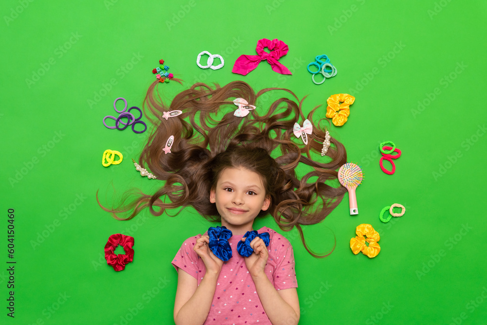 Children's hair accessories. A little girl with her hair down lies among the hairpins and elastic bands. Hairstyles for princesses. Green isolated background.