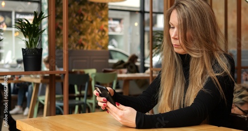 Fashionable woman sit alone in cafe and check news or social media on smartphone. Beautiful young lady with long brown hair in black turtleneck spend free time online in internet.