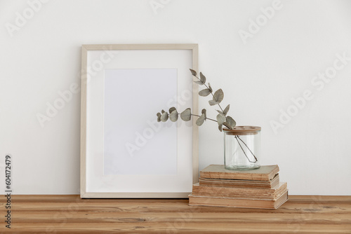  Empty wooden picture frame  poster mockup on white wall background. Glass vase with green eucalyptus tree branches on table and books. Neutral still life interior.Empty copy space.Modern art display