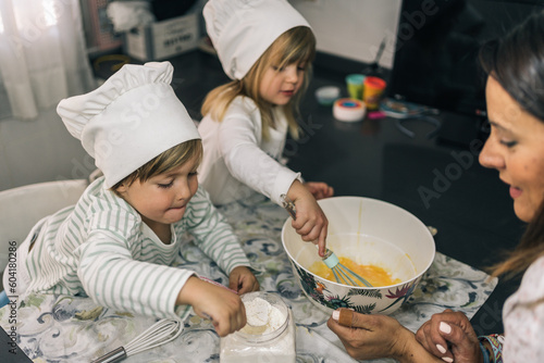 Pair of blond brother and sister children baking homemade cupcakes. One of them scoops flour with a spoon and the other one beats eggs.