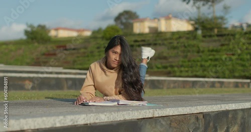 Young teenage Latina college student studying for an exam in an outdoor park with books