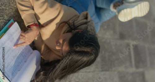 Young teenage Latina college student studying for an exam in an outdoor park with books, vertical shot