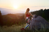 Woman tourist camping outdoors at sunset. Back view of slim woman near tent, admiring landscape in mountains. Female tourist wearing sportswear traveling in summer. Concept of beauty of nature.