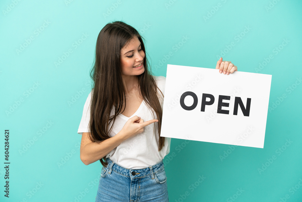 Young caucasian woman isolated on blue background holding a placard with text OPEN and pointing it Young caucasian woman isolated on blue background holding a placard with text OPEN and pointing it