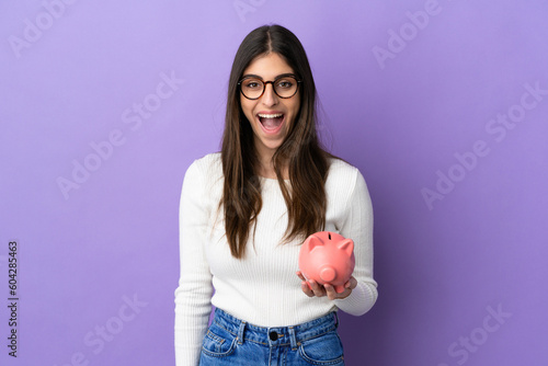 Young caucasian woman holding a piggybank isolated on purple background with surprise facial expression