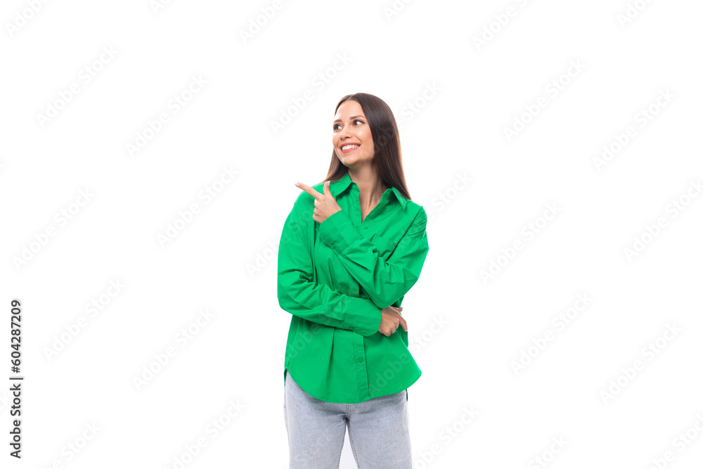 young european female model with well-groomed black hair dressed in a green shirt points her finger to the side on a white background with copy space young european female model with well-groomed black hair dressed in a green shirt points her finger to the side on a white background with copy space