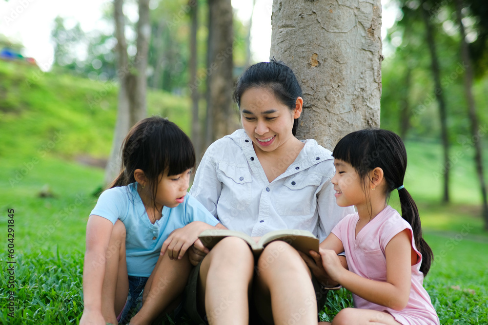Happy mother and two daughters having fun and enjoying reading at the park. Mother and daughter resting in the summer garden, she reads fairy tales to her daughter. Happy mother and two daughters having fun and enjoying reading at the park. Mother and daughter resting in the summer garden, she reads fairy tales to her daughter.