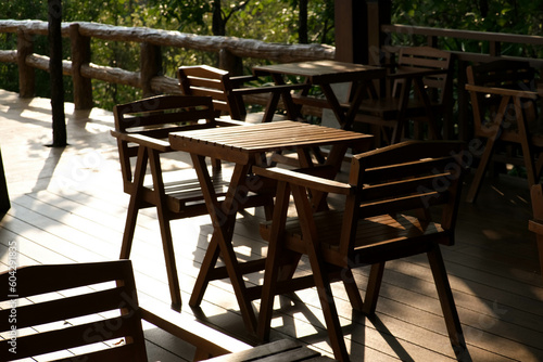 Wooden tables and chairs at outdoor cafe terrace in park. Empty garden furniture surrounded by green garden. © Pornpimon