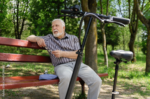 A positive, elderly man went for a walk to the park, stopped and sat down to rest on a bench next to an electric scooter. Summer, sunny day. Healthy lifestyle of the elderly. photo