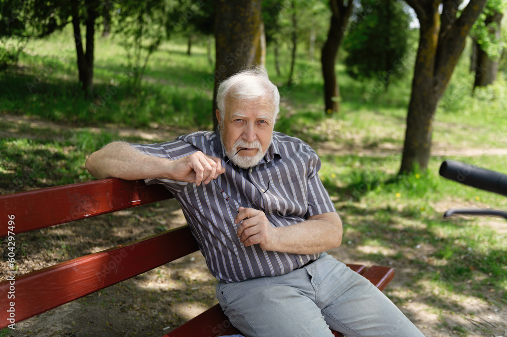 Fototapeta premium A positive, elderly man went for a walk to the park, stopped and sat down to rest on a bench next to an electric scooter. Summer, sunny day. Healthy lifestyle of the elderly.