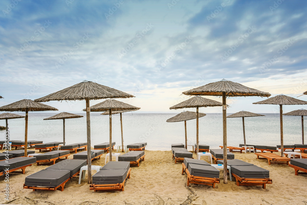 Wooden beach loungers with soft cushions and straw sun umbrellas right on the sea in a tourist resort in Greece against a cloudy blue sky, copy space