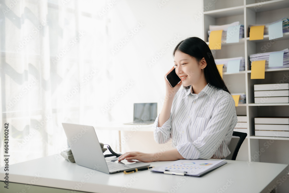 Business asian woman Talking on the phone and using a laptop with a smile while sitting in office Business asian woman Talking on the phone and using a laptop with a smile while sitting in office