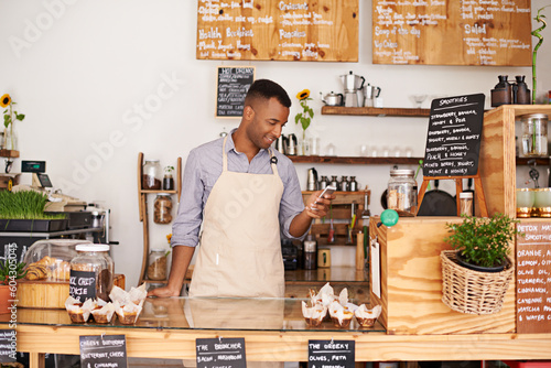 Black man, coffee shop and store phone of an entrepreneur with happiness from small business. Cafe, mobile and barista looking at online app with a smile at bakery and restaurant feeling happy