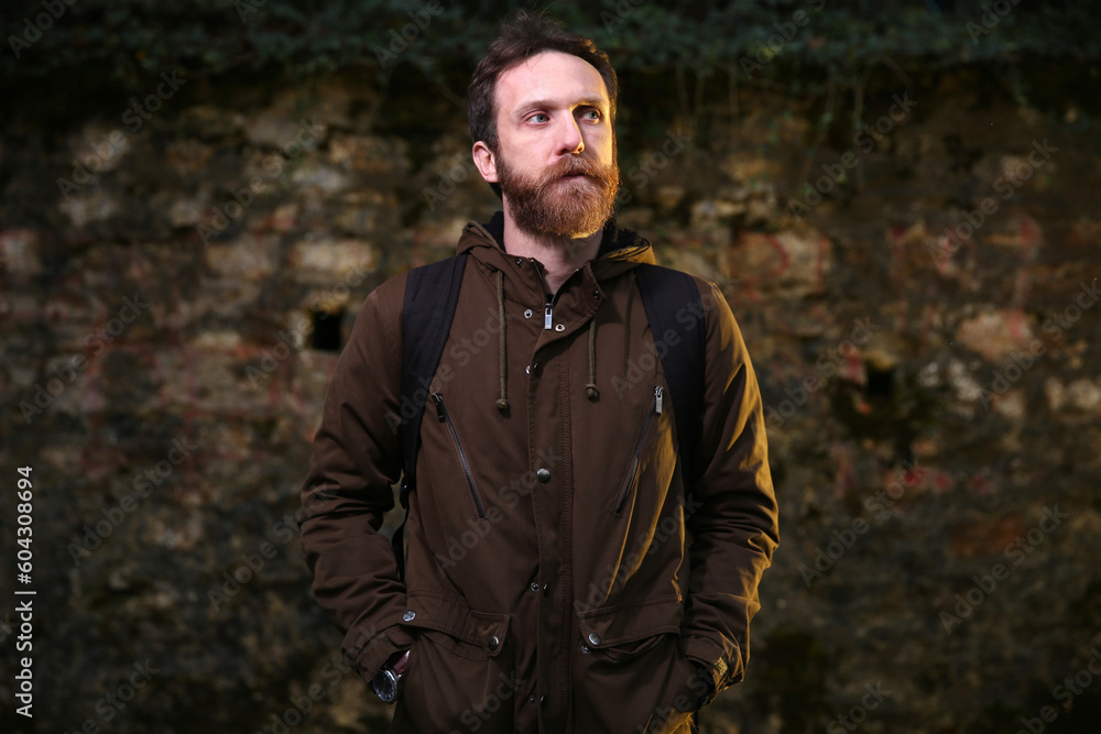 Trekking lover, adventurer, bearded Caucasian man with a brown jacket posing in front of a brick wall