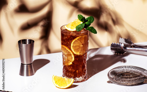 Long Island ice tea cocktail drink with vodka, rum, tequila, gin, liquor, lemon juice, cola and ice with lemon slice and mint in highball glass. Beige background, hard light, bar tools