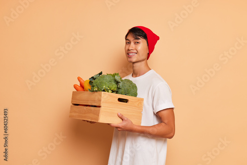 Cheerful Asian guy in white t-shirt and red hat standing inside the studio holding box of fresh vegetables, posing on background with kindly smile, organic food concept, copy space photo