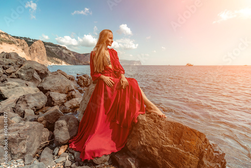 Red dress sea woman. A blonde with flowing hair in a long red dress sits on a rock near the sea. The concept of trips, a photo shoot at the sea