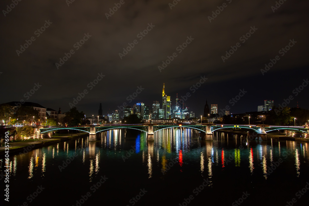 Cityscape  in Frankfurt, Germany featuring a bridge, river, nighttime city illumination reflection, and a downtown skyscrapers background.