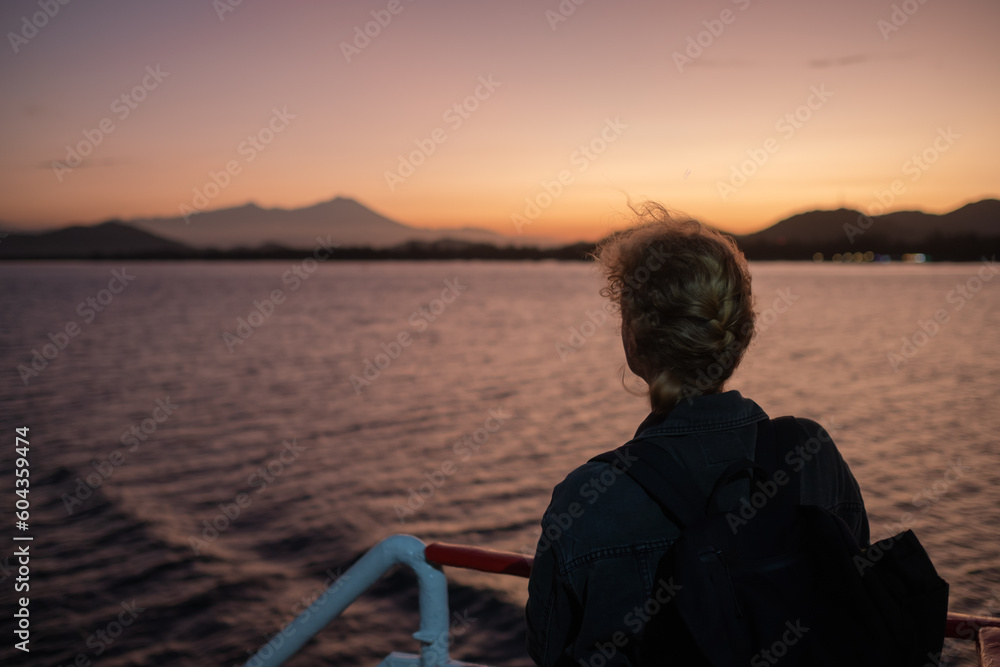 Fototapeta premium Young Blonde Woman with a Backpack Traveling on a Ferry, Admiring a Stunning Ocean Sunrise
