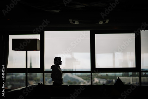 Silhouette of young cute girl who pressed against the glass and admiring the panoramic view of city from the tall building. Selective focus on hand. Blurred background.