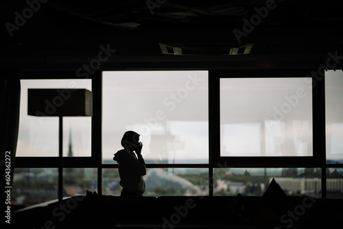 Silhouette of young cute girl who pressed against the glass and admiring the panoramic view of city from the tall building. Selective focus on hand. Blurred background.