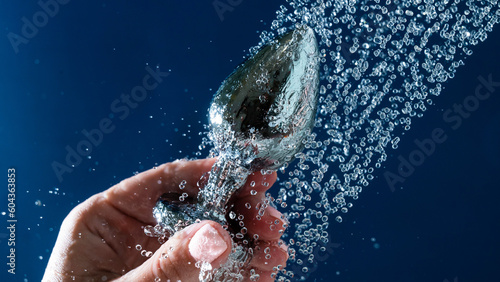 Woman washing silver anal plug under shower on blue background. 
