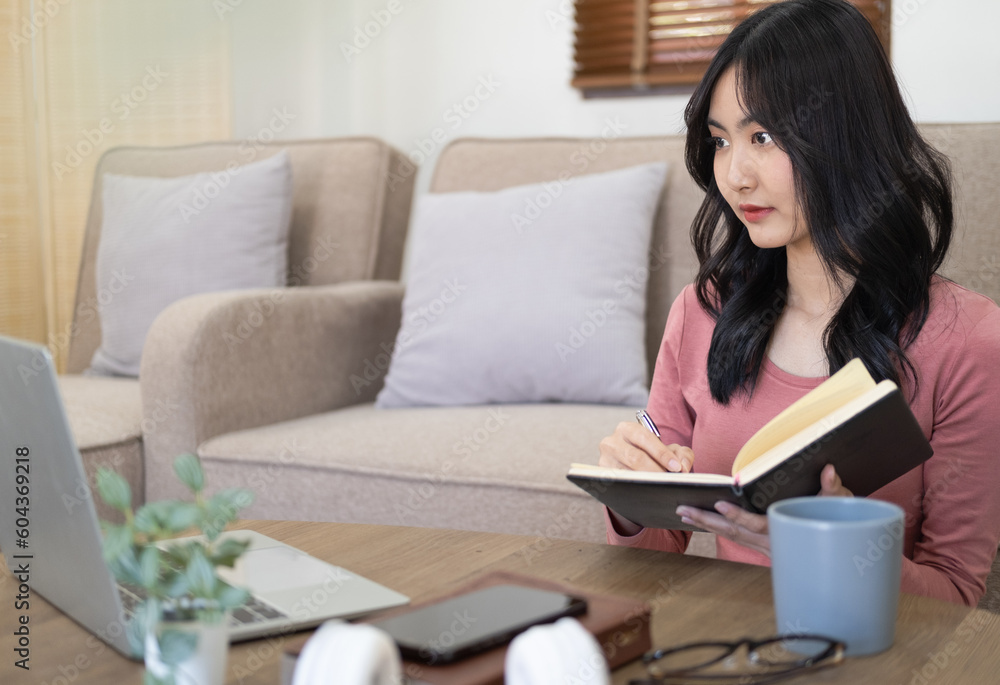 Modern Asian female student in stylish casual clothes, studying remotely using a laptop, listening to an online lesson, taking notes in a notebook, while sitting in a living room. Online education Modern Asian female student in stylish casual clothes, studying remotely using a laptop, listening to an online lesson, taking notes in a notebook, while sitting in a living room. Online education