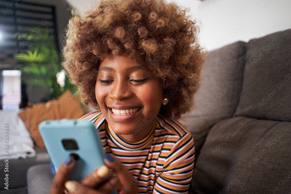 Close up of smiling African American pretty woman using cell phone lying on sofa in living room. Happy young black afro girl flirting on mobile dating app at home. People looking for life partner. Close up of smiling African American pretty woman using cell phone lying on sofa in living room. Happy young black afro girl flirting on mobile dating app at home. People looking for life partner.