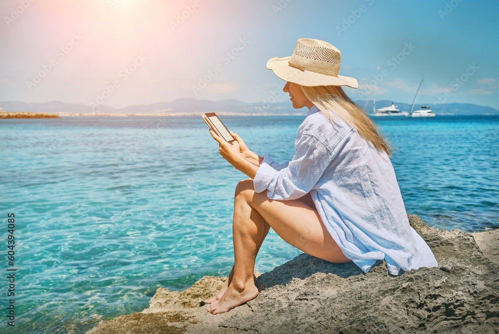 Happy woman outdoors lifestyle watching, reading on tablet ebook on the beach in summer day. Wearing wide brimmed hat, with uv protection. Concept of beach vacation.