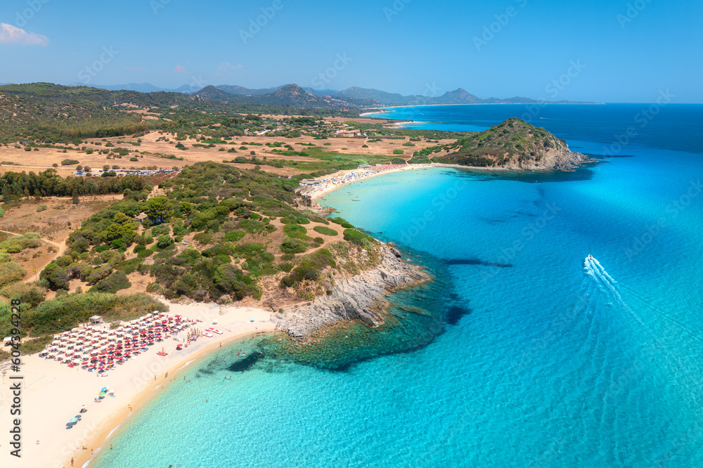 Aerial view of white sandy beach, umbrellas, mountains, green trees, yacht in blue sea at sunset in summer. Tropical landscape. Travel in Sardinia, Italy. Top view of ocean with transparent water