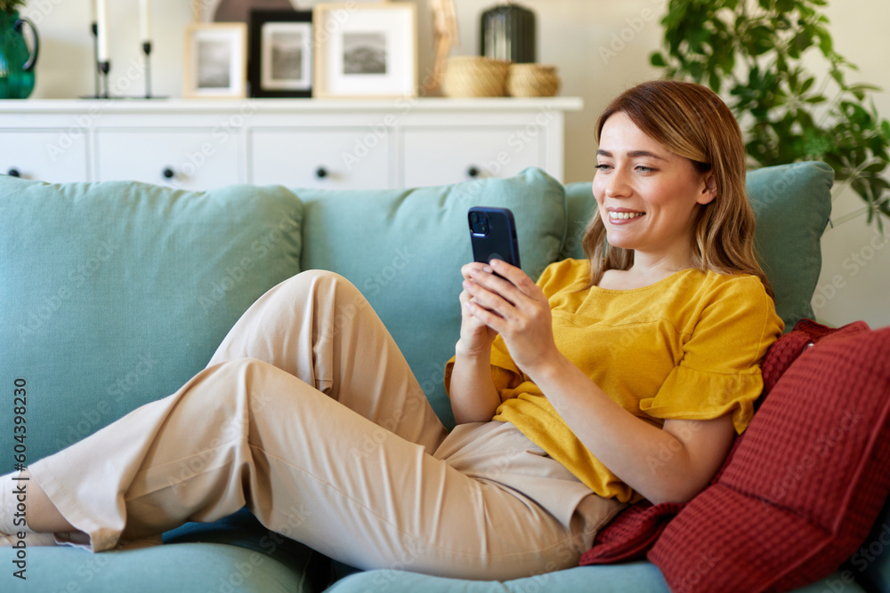 Happy woman text messaging on mobile phone while relaxing in the living room at home Happy woman text messaging on mobile phone while relaxing in the living room at home