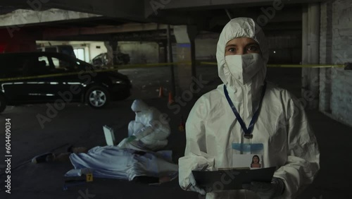 Medium portrait shot of woman in white protective suit and face mask standing at murder scene, writing on clipboard, then looking at camera, and colleague examining corpse of victim in background photo