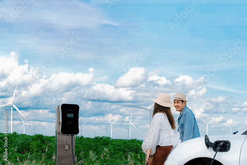 Concept of progressive happy young couple enjoying their time at windmill farm with electric vehicle. EV car driven by clean renewable energy from wind turbine generator for charging station. photo