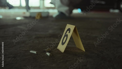 Close-up shot of yellow police marker with number 6 on ground next to cigarette stubs at urban crime scene, and unrecognizable forensic experts in white suits collecting evidence in background photo