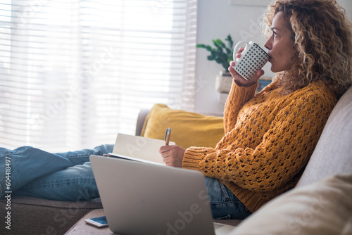 One young adult woman having relax at home drinking from cup and writing no notes with laptop on the sofa near her Fototapet