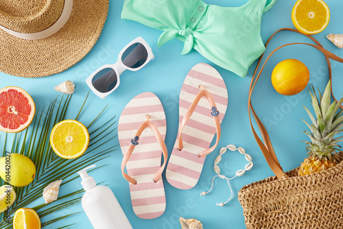 Summer getaway concept. Top view flat lay of stylish bag, trendy sunhat, swimsuit, flip flops, sunscreen bottle, seashells, orange fruit slices, pineapple and palm leaf on light blue background