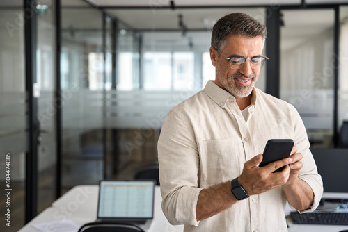 Smiling mature Latin or Indian businessman holding smartphone in office. Middle aged manager using cell phone mobile app. Digital technology application and solutions for business. Copy space