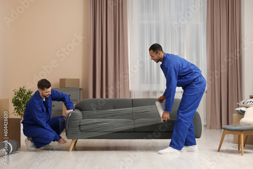Male movers with stretch film wrapping sofa in new house photo