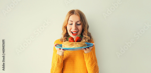 Cooking, portrait of happy laughing cheerful young woman housewife holding plate with stack of pancakes with strawberries on gray wall background