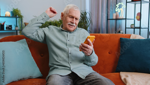 Sad displeased senior grandfather man use smartphone typing browsing, loses becoming surprised sudden lottery results, bad news, fortune loss, fail, deadline, virus. Elderly guy at home room on couch