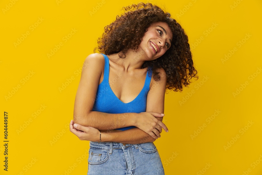 Woman with curly afro hair in a blue t-shirt on. yellow background signs with her hands, look into the camera, smile with teeth and happiness, copy space Woman with curly afro hair in a blue t-shirt on. yellow background signs with her hands, look into the camera, smile with teeth and happiness, copy space