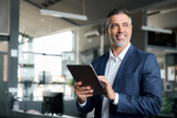 Happy middle aged business man ceo wearing suit standing in office using digital tablet. Smiling mature businessman professional executive manager looking away thinking working on tech device.