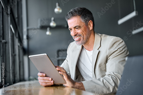 Happy mid aged business man wearing suit standing in modern office using digital tablet. Mature businessman professional manager holding tab working on financial data on fintech device.
