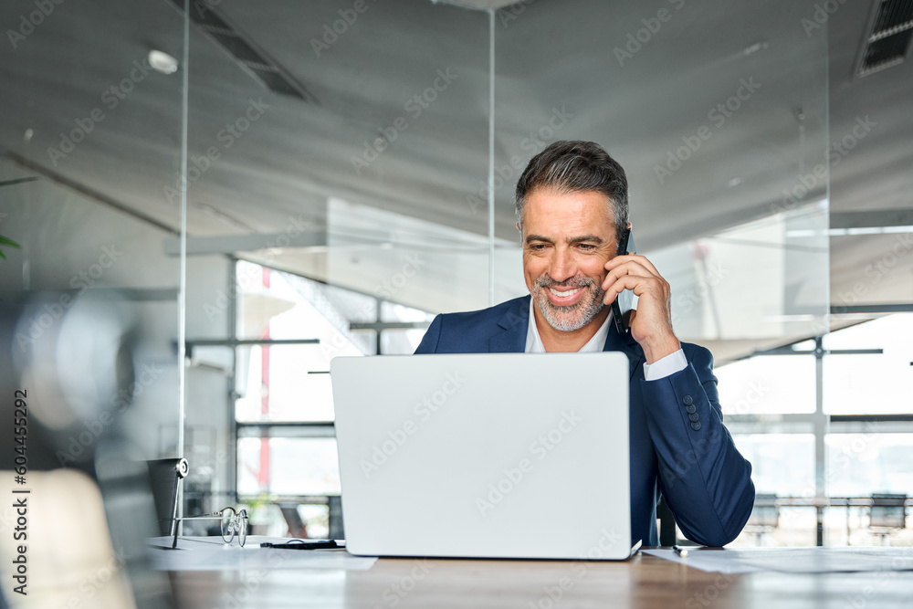 Fototapeta premium Happy middle aged business man talking on mobile phone working on laptop in office. Professional businessman executive wearing suit making call on cell using computer communicating with client.