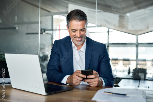 Photographie Happy mid aged business man ceo executive, financial expert investor wearing suit sitting in modern office holding smartphone using mobile phone managing digital tech work transactions at workplace