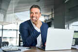 Happy smiling middle aged professional business man company executive ceo manager wearing suit sitting at desk in office working on laptop computer laughing at workplace. Authentic candid photo.