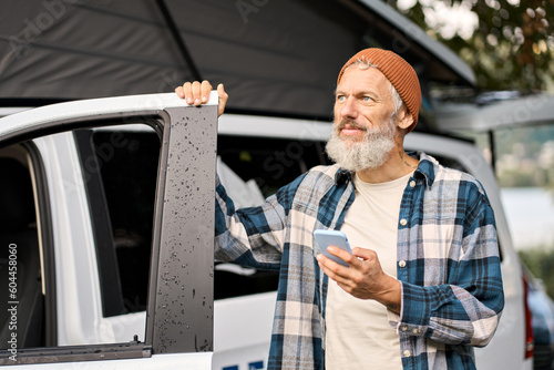 Older man standing near rv camper van on vacation using mobile phone. Elder mature active traveler holding smartphone in camping tourism nature park advertising campervan booking rental service. photo