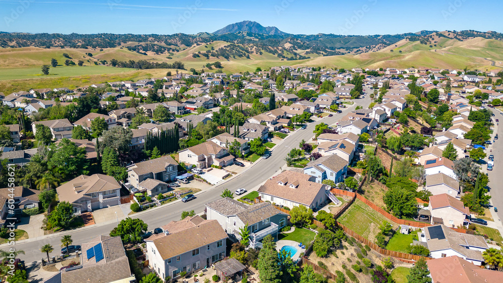 .A breathtaking aerial view of Antioch, California: houses nestled amidst verdant hills, crisscrossed by meandering streets, all under a picturesque blue sky.