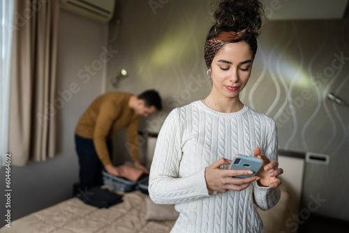 couple young beautiful woman and man in hotel room wife or girlfriend use mobile phone for texting sens sms messages while husband or boyfriend is unpacking or packing baggage on the bed copy space