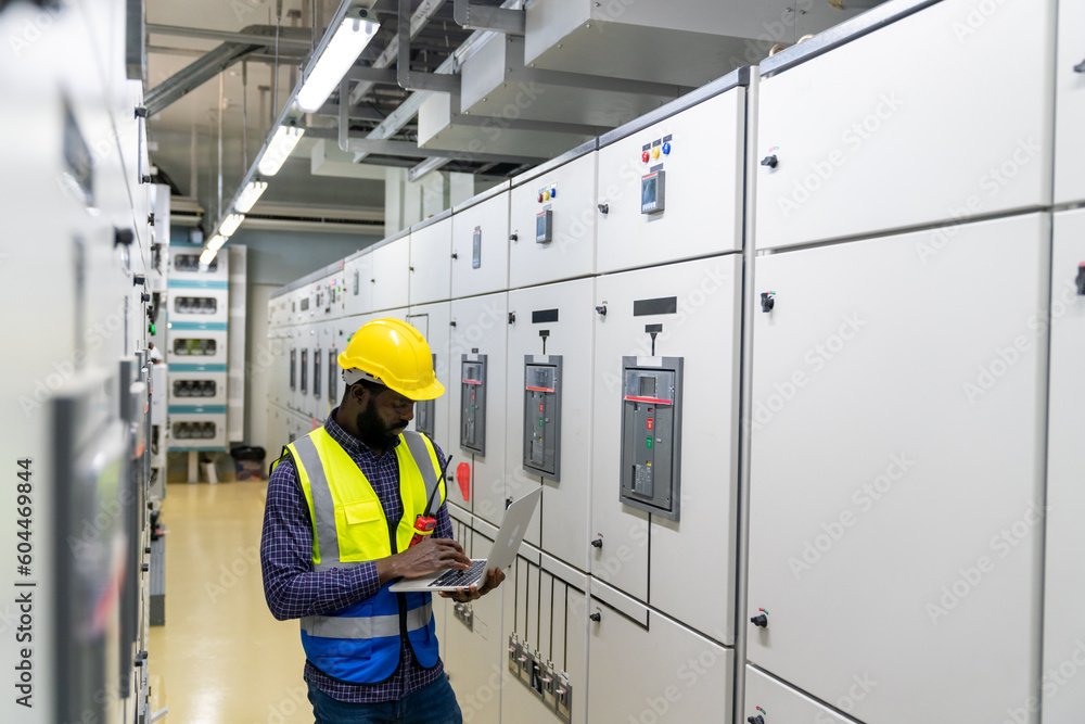 Professional African male engineer in safety uniform working at factory server electric control panel room. Industrial technician worker maintenance checking power system at manufacturing plant room. Professional African male engineer in safety uniform working at factory server electric control panel room. Industrial technician worker maintenance checking power system at manufacturing plant room.